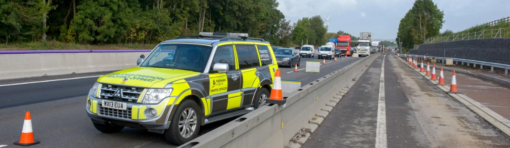Highway maintenance vehicle parked by traffic cones on a divided multi-lane road, supporting emergency response efforts with moving vehicles and roadwork barriers visible.