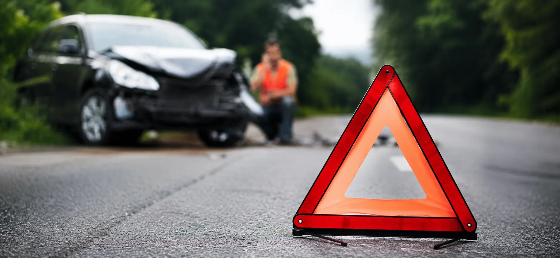 A red warning triangle is placed on a road near a damaged car with a person in a safety vest in the background, emphasizing important measures that can help save lives after a crash.